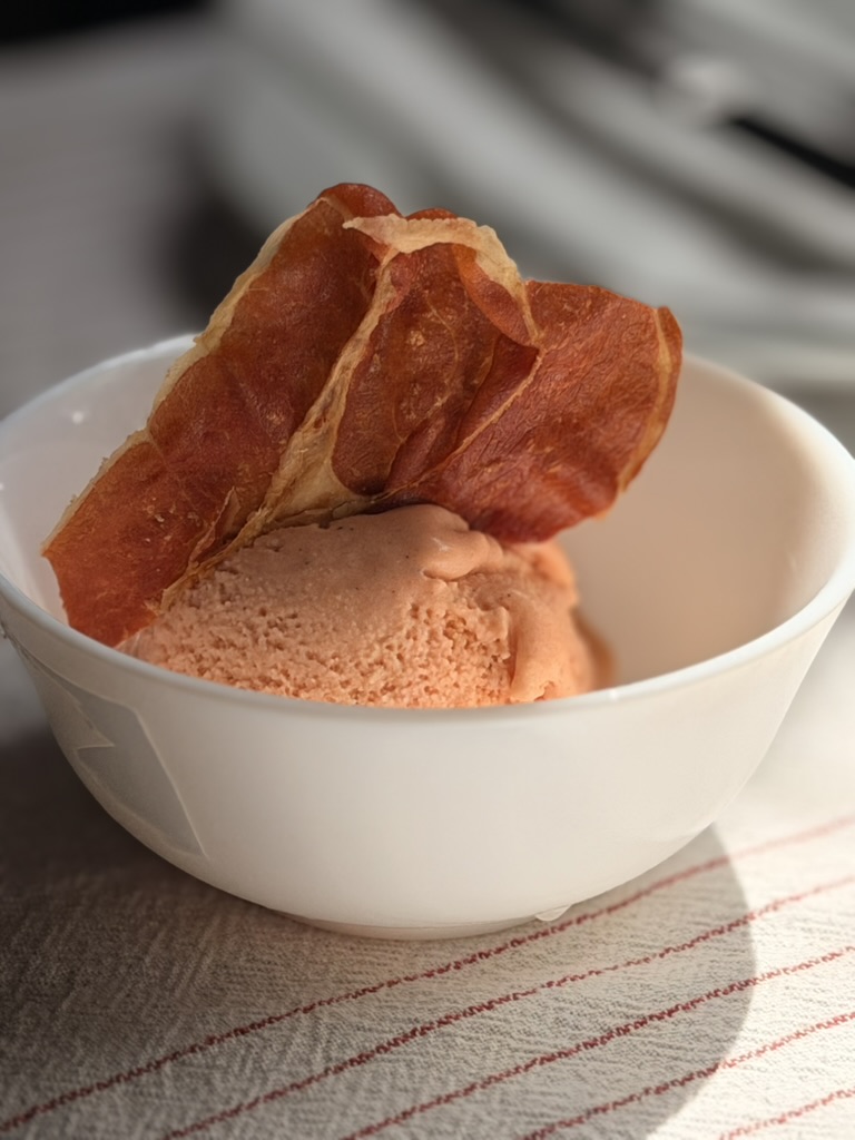 A scoop of pale coral gazpacho ice cream in a white bowl, topped with a crisp shard of Serrano ham