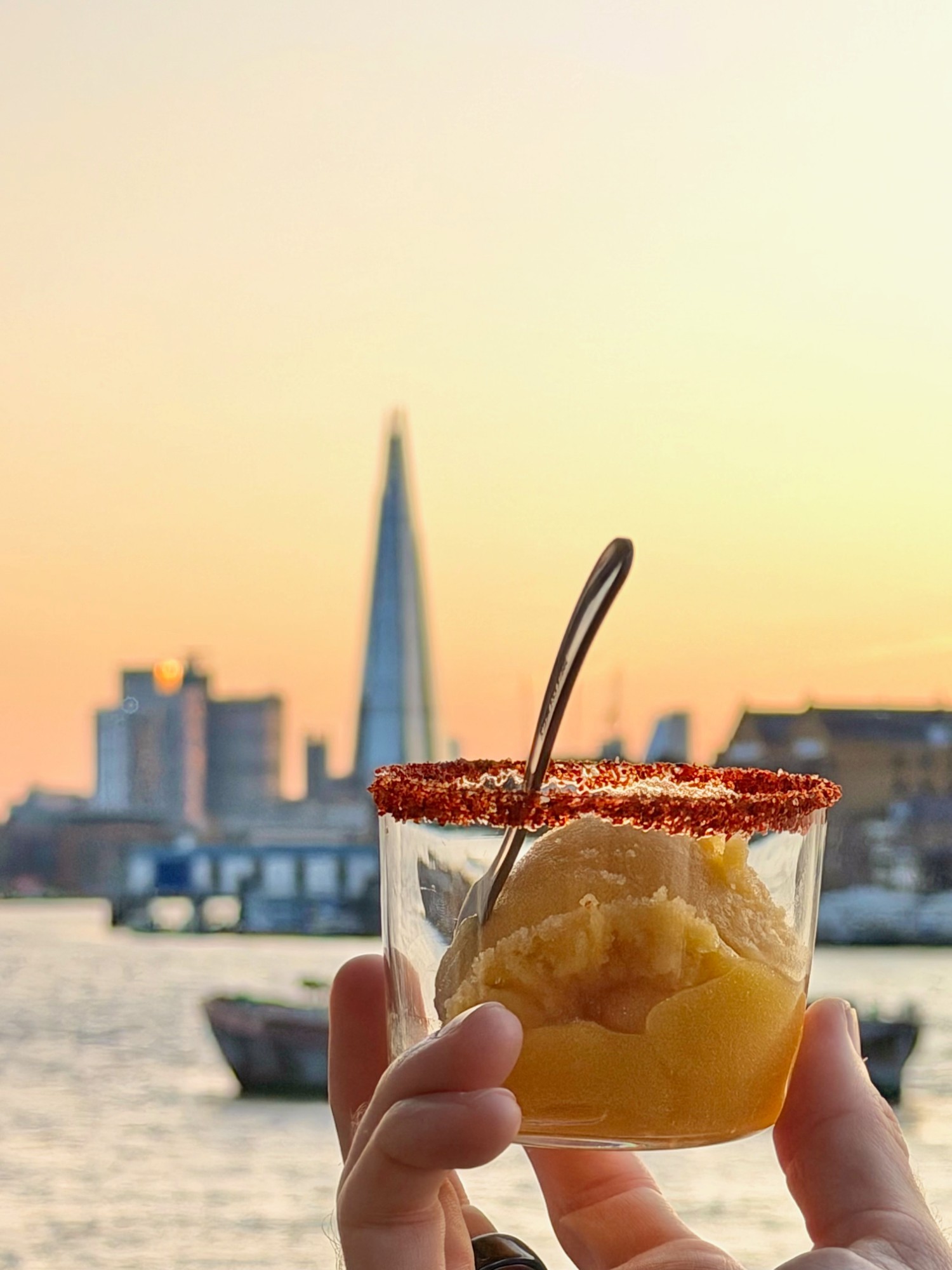 A hand holding a mango margarita in a chilli-salt-rimmed glass against a London sunset, the Shard in the background